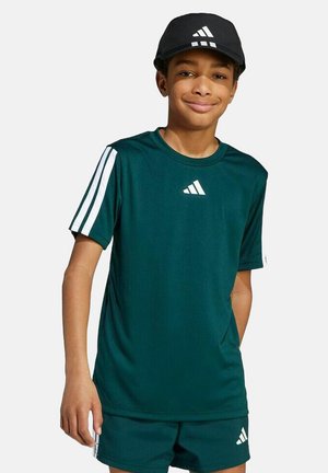 Junge lächelt, trägt dunkelgrüne Adidas-Sportbekleidung mit weißen Streifen und eine schwarze Adidas-Kappe, steht vor einem schlichten Hintergrund.