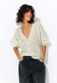 Cardigan léger de couleur crème en tricot, avec un col en V profond, des fermetures à boutons et des motifs ajourés complexes ; associé à un jean noir.
