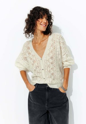Cardigan léger de couleur crème en tricot, avec un col en V profond, des fermetures à boutons et des motifs ajourés complexes ; associé à un jean noir.