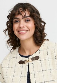 Jakna iz volnene mešanice v kremni barvi z mrežnim vzorcem, s krožno ovratnico in rjavimi usnjenimi detajli pri ovratniku.