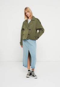 Oliivinvihreä, lyhyt kaksoisrintainen takki isoilla napeilla, yhdistetty vaaleansiniseen denimihameeseen, jossa on edessä halkio, ja mustiin tennareihin.