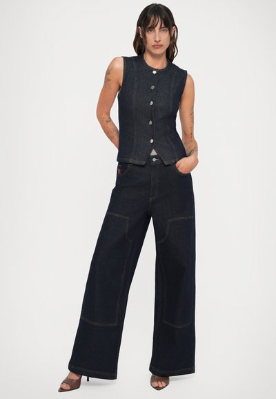 Γυναίκα με τατουάζ που φοράει αμάνικο σκούρο τζην γιλέκο και φαρδιές τζιν παντελόνες, στέκεται με το ένα χέρι στην τσέπη και το άλλο πίσω από την πλάτη.