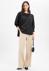 Czarna satynowa bluzka z szerokimi, marszczonymi rękawami, zestawiona z beżowymi, szerokimi spodniami oraz czarnymi szpilkami z ostrym noskiem, trzyma małą czarną torebkę.
