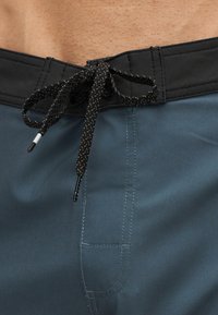 Κοντινή λήψη δεμένου μαύρου κορδονιού σε μπλε μαγιό με μαύρη μέση που φοριέται από ένα άτομο.