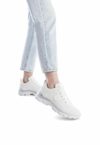 Zapatillas de punto blancas con una parte superior texturizada, cierre de cordones en la parte delantera y un acento de talón de plástico transparente; combinadas con jeans azul claro con vuelta.