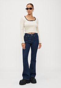 White long-sleeve crop top with black trim and ruffled edges, paired with navy blue flared jeans and black platform shoes. Sunglasses worn.