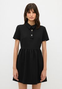 Vestido negro de camisa con cuello, mangas cortas, tres botones a presión plateados y falda con frunzidos, hecho de una tela suave.