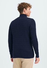 Mornarsko pleteni pulover s turtleneck ovratnikom z teksturiranim kabelskim vzorcem, rebrastimi manšetami in robom; oblečen z bež kordovnimi hlačami.