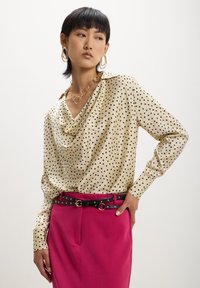 Blouse beige à pois avec un col drapé et des manches longues, associée à une jupe crayon rose avec une ceinture noire et des accents dorés.