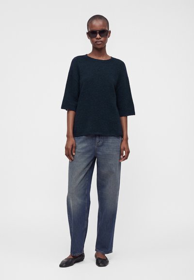 Femme portant des lunettes de soleil foncées, un pull noir ample à manches courtes, un jean bleu délavé et des chaussures plates noires, debout devant un fond uni.