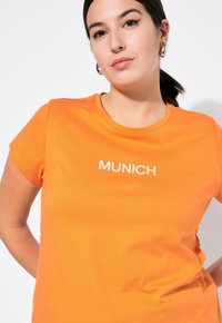 Oranges Baumwoll-T-Shirt mit rundem Ausschnitt und kurzen Ärmeln, versehen mit dem gestickten Schriftzug "MÜNCHEN" in Weiß auf der Vorderseite.