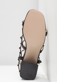 Semelle d'une sandale noire à talon haut carré avec des brides cloutées, présentée sur fond blanc.