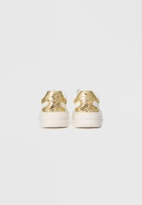 Weiße Sneaker mit dicken Sohlen und mit goldenen Pailletten überzogenen Fersentabs, von hinten auf einem schlichten hellgrauen Hintergrund gezeigt.