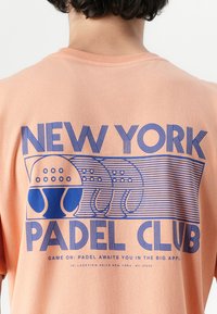 Persikinio spalvos marškiniai su mėlynu piešiniu ir užrašu "NEW YORK PADEL CLUB", taip pat iliustracijomis apie padelio raketes ir kamuoliukus.