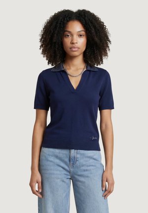 Femme aux cheveux bouclés portant un haut bleu marine à manches courtes avec un col à motifs, un collier chaîne argenté et un jean bleu clair.