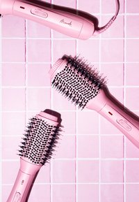 Ensemble d'outils de coiffure rose comprenant une brosse ronde avec des poils et un manche, dotée de boutons et d'une prise texturée, disposée sur une surface carrelée rose.