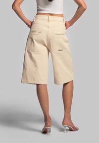 Pantaloni corti beige fino al ginocchio realizzati in tessuto testurizzato, dotati di due tasche posteriori e marchio ricamato in stile corsivo.