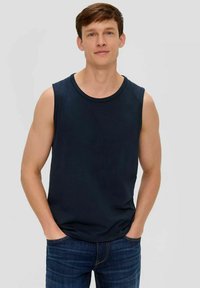 Junger Mann mit kurzen braunen Haaren, der ein ärmelloses Marinehemd und eine blaue Jeans trägt, und mit den Händen in den Taschen vor einem schlichten Hintergrund steht.