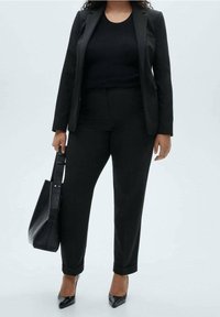 Costume noir sur mesure avec blazer cintré et pantalon, haut noir côtelé, et talons noirs pointus. Comprend un sac noir structuré avec une sangle.