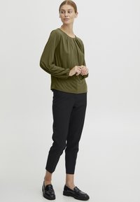 Oliven grønn genser med lange ermer og rund hals, med rynkede detaljer, kombinert med tettsittende svarte bukser og blanke svarte loafers.