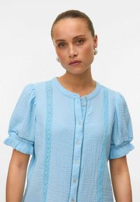 Camicia azzurra chiara con maniche corte a sbuffo, tessuto testurizzato, dettagli in pizzo lungo la parte anteriore e chiusura con bottoni e scollatura rotonda.