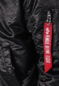 Casaco bomber preto com um padrão de estrelas subtil, bolsos laterais e uma etiqueta vermelha "Remover Antes do Voo" presa com um clip metálico.