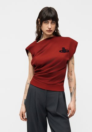 Sötét hajú nő piros ujjatlan felsőben, amelyen logó látható, és szürke pliszírozott nadrágban, mindkét karján tetoválásokkal, egyszínű háttér előtt.