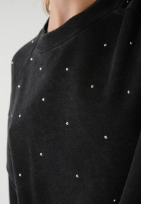 Felpa nera con scollo rotondo, decorata con strass sparsi su tutta la superficie. Il materiale sembra morbido e testurizzato.