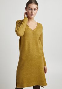 Robe en laine moutarde à manches longues avec un décolleté en V, fentes sur les côtés et une finition texturée. Accentuée d'une petite étiquette logo en bas.