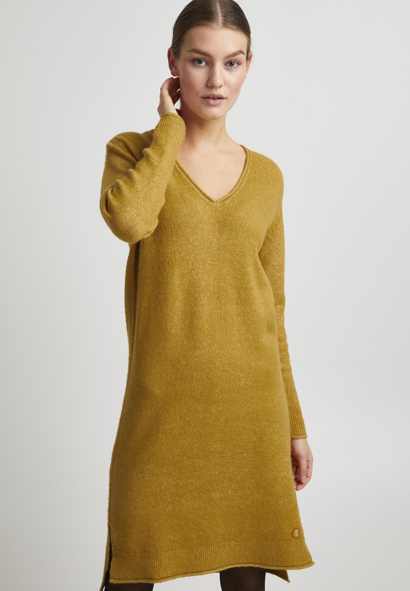Robe en laine moutarde à manches longues avec un décolleté en V, fentes sur les côtés et une finition texturée. Accentuée d'une petite étiquette logo en bas.