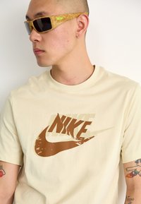 Kreminė marškinė su ryškiai ruduo Nike logotipu, derinami su gintaro spalvos akinių nuo saulės, turinčių juodus stiklus ir žalius akcentus.