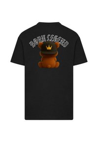 Mustaga T-särk, millel on prinditud pruun karu musta mütsiga, millel on kroon, ja tekst "SÜNDINUD LEGEND" karu kohal.