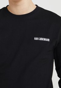 Svart långärmad tröja i bomull med rund halsringning och en liten vit broderad logotyp som säger "HAN KJØBENHAVN" på bröstet.