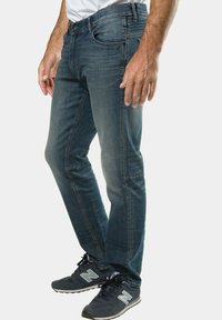 Jeans slim ajustados para hombre en denim azul oscuro, con un diseño clásico de cinco bolsillos y detalles de costura, combinados con zapatillas deportivas negras.