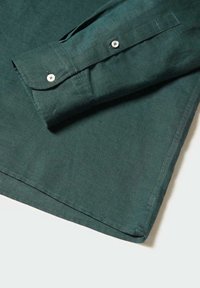 Polsino di una camicia verde a maniche lunghe con due bottoni bianchi e cuciture visibili su uno sfondo chiaro.