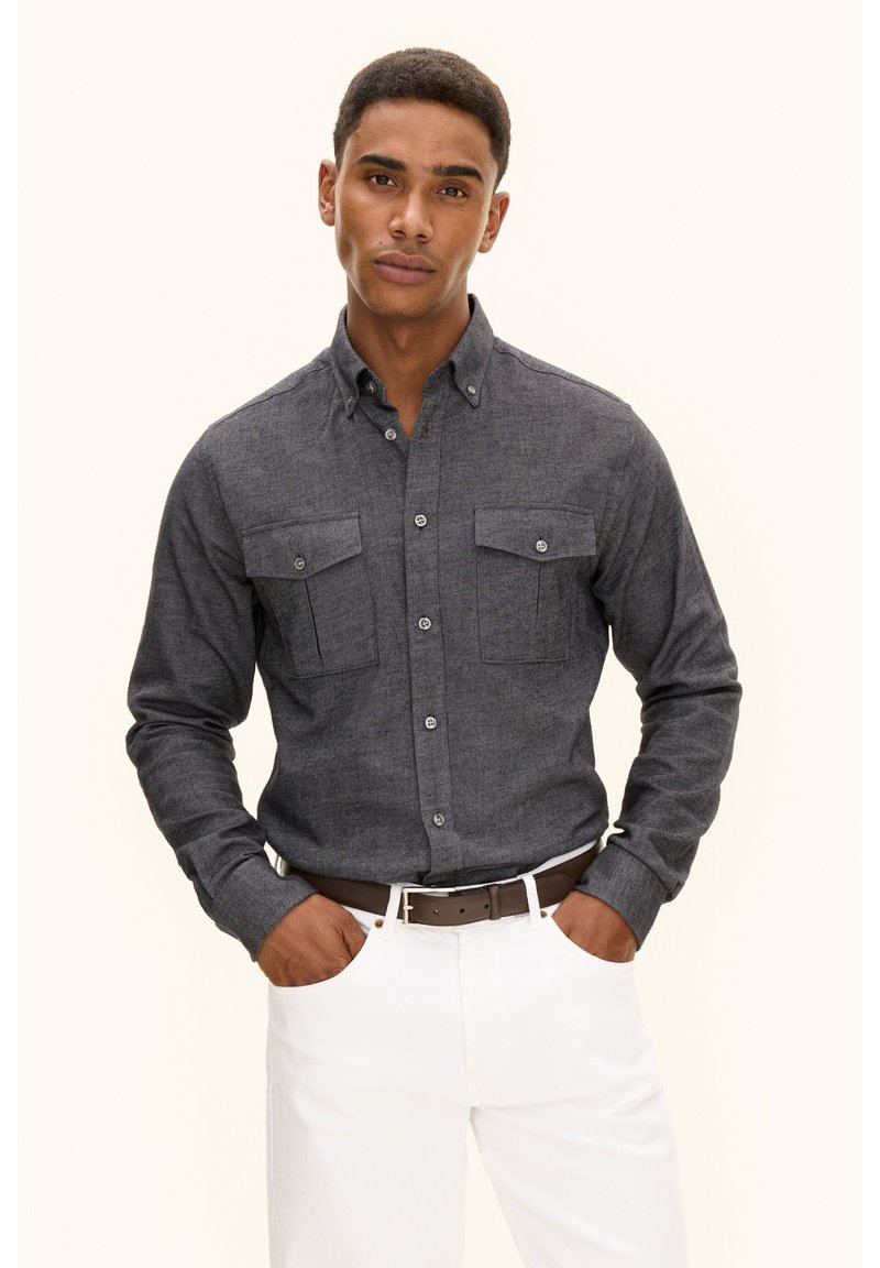 Camicia grigia scura da uomo con bottoni, due tasche sul petto e colletto, realizzata in tessuto texturizzato. Abbinata a pantaloni bianchi e una cintura marrone.