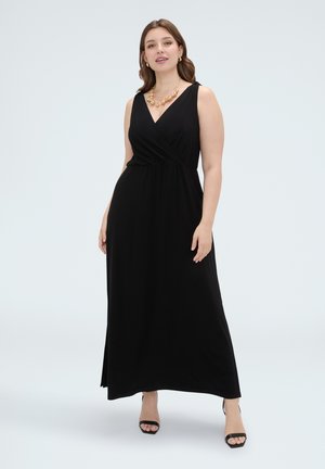 Woman wearing a sleeveless black maxi dress with a V-neck, gold necklace, hoop earrings, and black open-toe heels standing on a white background.