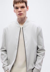 Ανοιχτό γκρι μπουφάν τύπου bomber με όρθιο γιακά, φερμουάρ μπροστά και ριμπ μανσέτες. Φορεμένο πάνω από μπεζ, υφασμένο πουλόβερ με υφή.