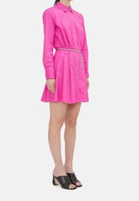 Abito fucsia con bottoni, maniche lunghe, vita cintrata e gonna a ruota. Presenta una cintura in catena argentata. Abbinato a mules con tacco a blocco neri.