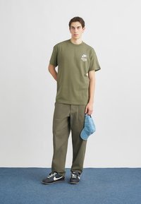 Λαδί βαμβακερό μπλουζάκι με μικρό λογότυπο, συνδυασμένο με φαρδιά λαδί παντελόνια και ανοιχτό μπλε καπέλο, με ουδέτερο φόντο.