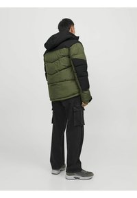 Giacca a vento in verde oliva e nero, con cappuccio, design trapuntato, tessuto testurizzato e polsini regolabili. Abbinata a pantaloni cargo neri.