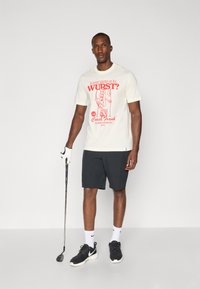 Short-sleeve cream t-shirt featuring a red graphic print, paired with dark shorts and black athletic shoes. Standing with a golf club.