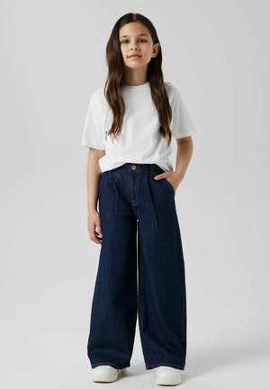 Camiseta blanca de manga corta combinada con pantalones vaqueros de pierna ancha en denim azul oscuro, con cierre de botón y bolsillos laterales. Zapatillas blancas completan el look.