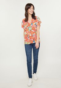 Blusa floreale con maniche corte, caratterizzata da un scollo a V. Fiori colorati in rosa, arancio e blu su uno sfondo crema. Abbinata a jeans blu e sneakers bianche.
