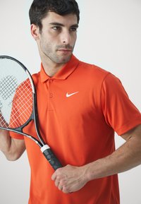 Oransje polo skjorte med krage og to knapper, med en hvit Nike-logo. Holder en tenniskølle med svart grep.