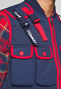 Gros plan sur une personne portant un gilet matelassé bleu marine avec bordure rouge, deux poches avant, boutons rouges, bretelles réglables noires et rouges, et une chemise à carreaux.