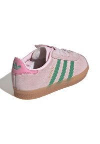 Pink suede sneaker with three green stripes, white laces, and a gum sole. Gold "GAZELLE" branding on the heel. Rounded toe design.