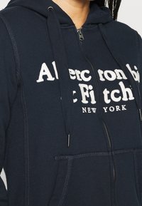 Marineblauwe zip-up hoodie van zachte stof, met witte tekst "Abercrombie & Fitch" en "NEW YORK", met trekkoorden en voorzakken.