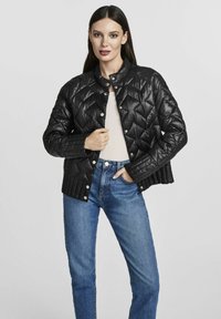Pikowana czarna kurtka puchowa z okrągłym kołnierzem, zapinana na napy, o teksturowanej tkaninie. Nosi ją się na jasną bluzkę, zestawioną z niebieskimi dżinsami.