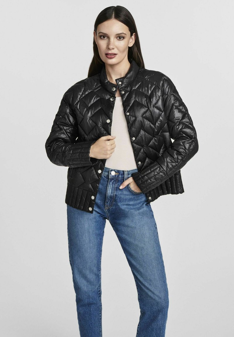 Giacca trapuntata nera con colletto tondo, chiusura a bottoni automatici e tessuto testurizzato. Indossata sopra a un top chiaro, abbinata a jeans blu.